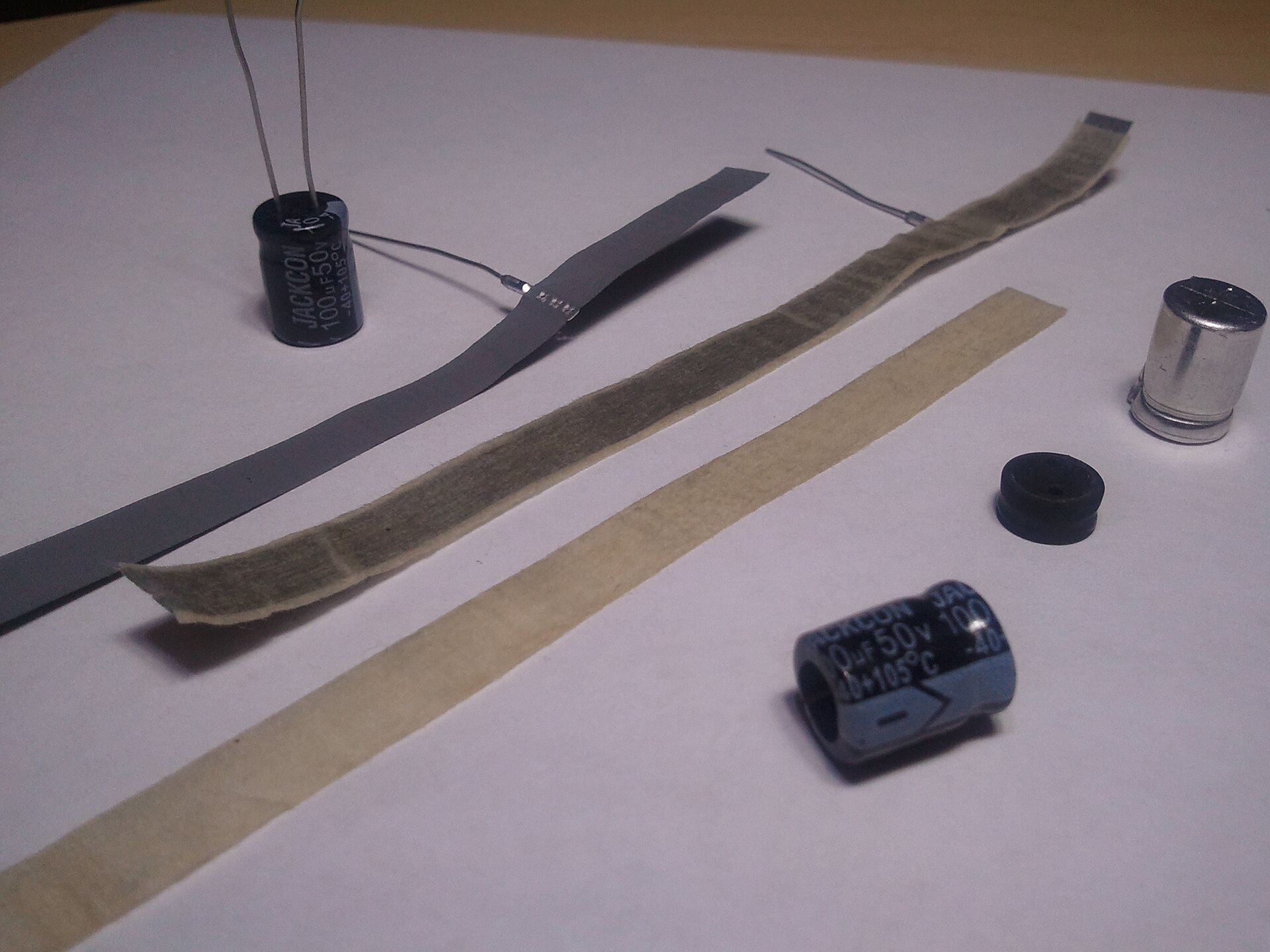 Figure 7 — A disassembled 100 µF aluminum electrolytic capacitor. The tall can unrolls into a long strip: two aluminum foils separated by an electrolyte-soaked paper spacer, wound in a spiral to pa…