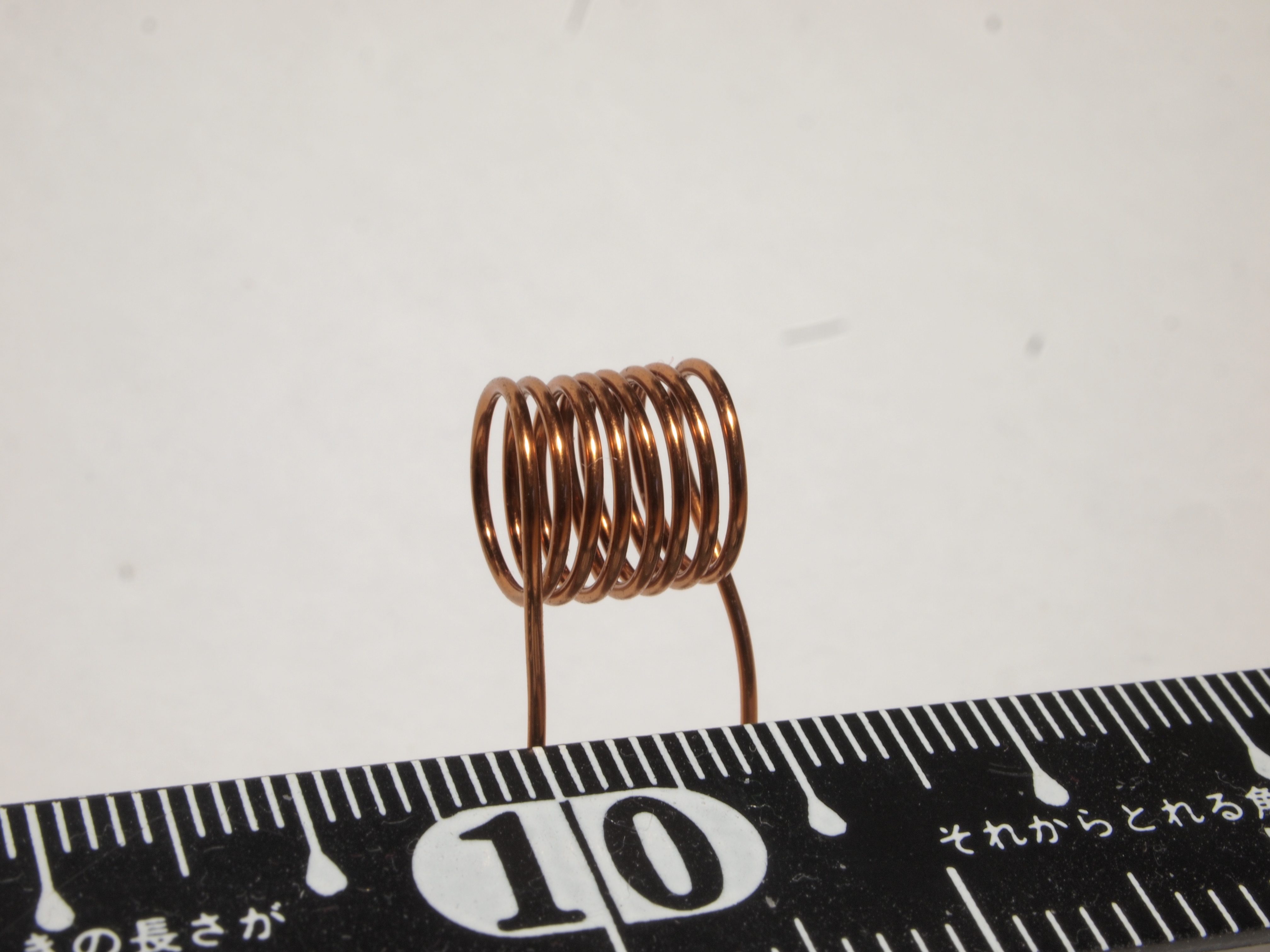 Figure 6 — A hand-wound single-layer air-core RF coil, self-supporting copper against a centimetre scale. No core, no former left in place: just wire, air, and geometry. Coils like this set the fre…