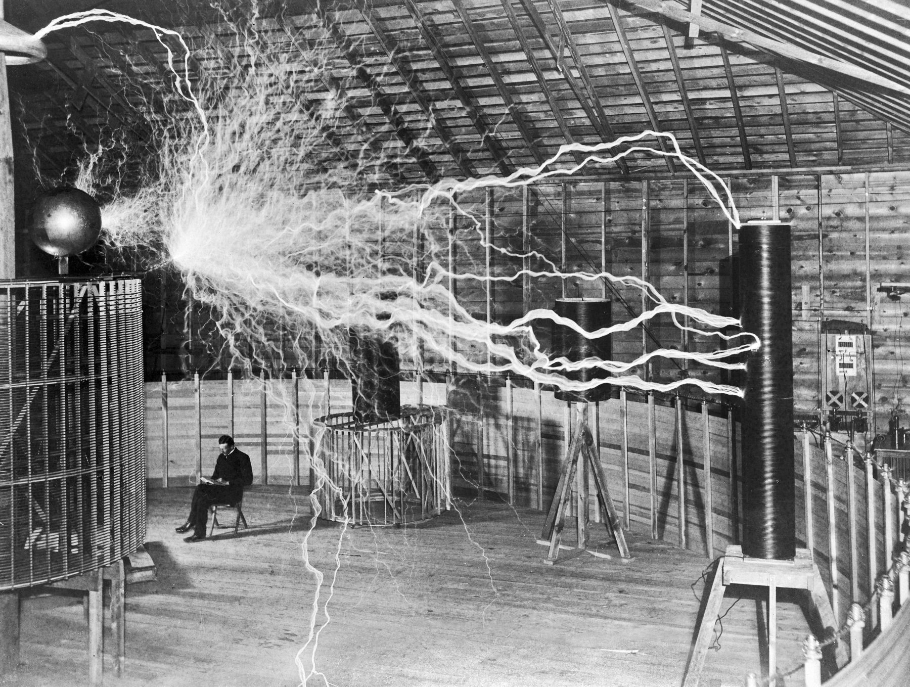 Figure 6 — Nikola Tesla in his Colorado Springs laboratory, 1899, seated beside a "magnifying transmitter." The dramatic sparks are a resonant air-core transformer at work — coils tuned to the same…