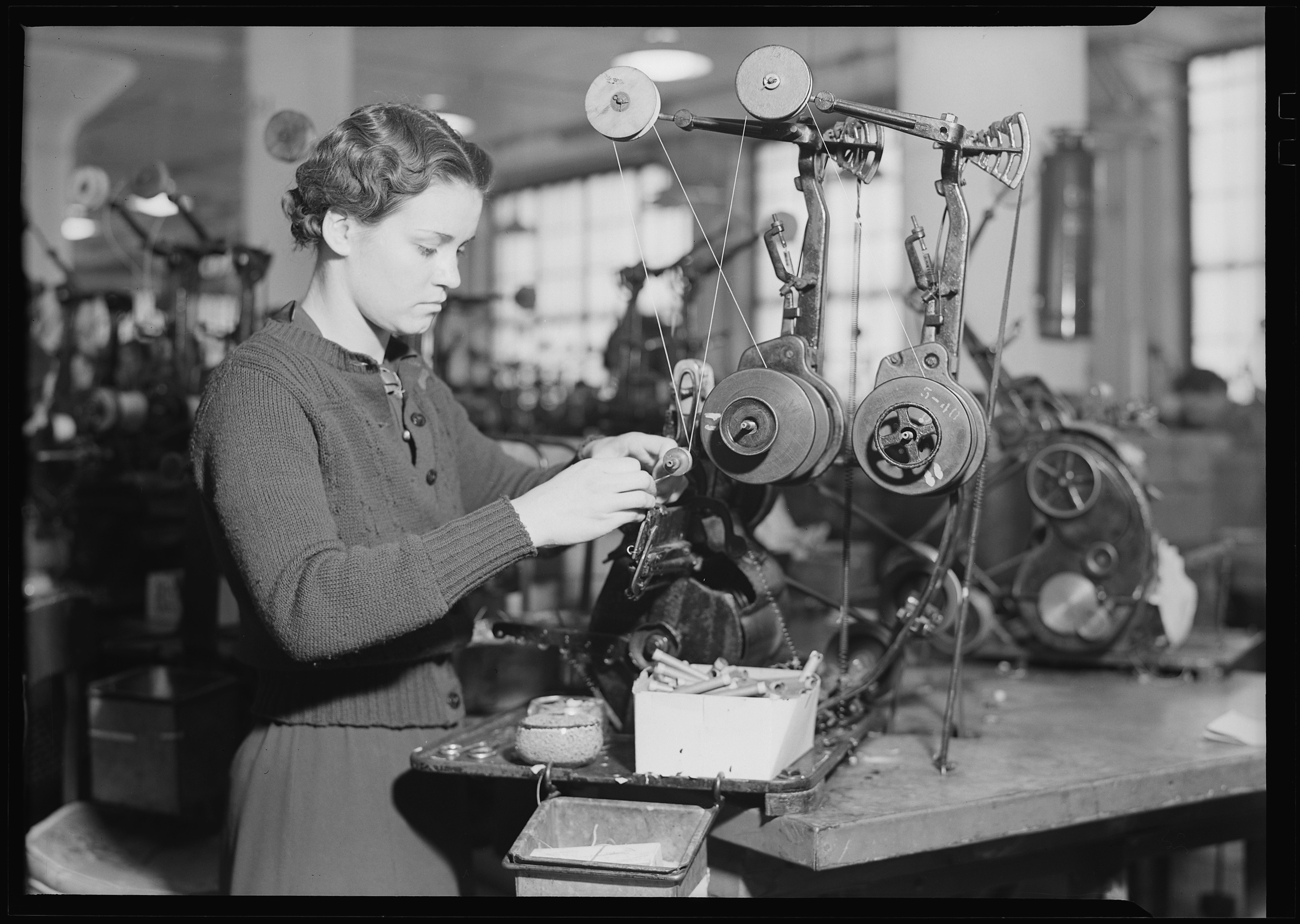 Figure 6 — A 1930s bench coil-winding machine in use. The wire runs up from the supply, over the two large felt-faced tension pulleys at right, down to the small forms the operator is winding; the …