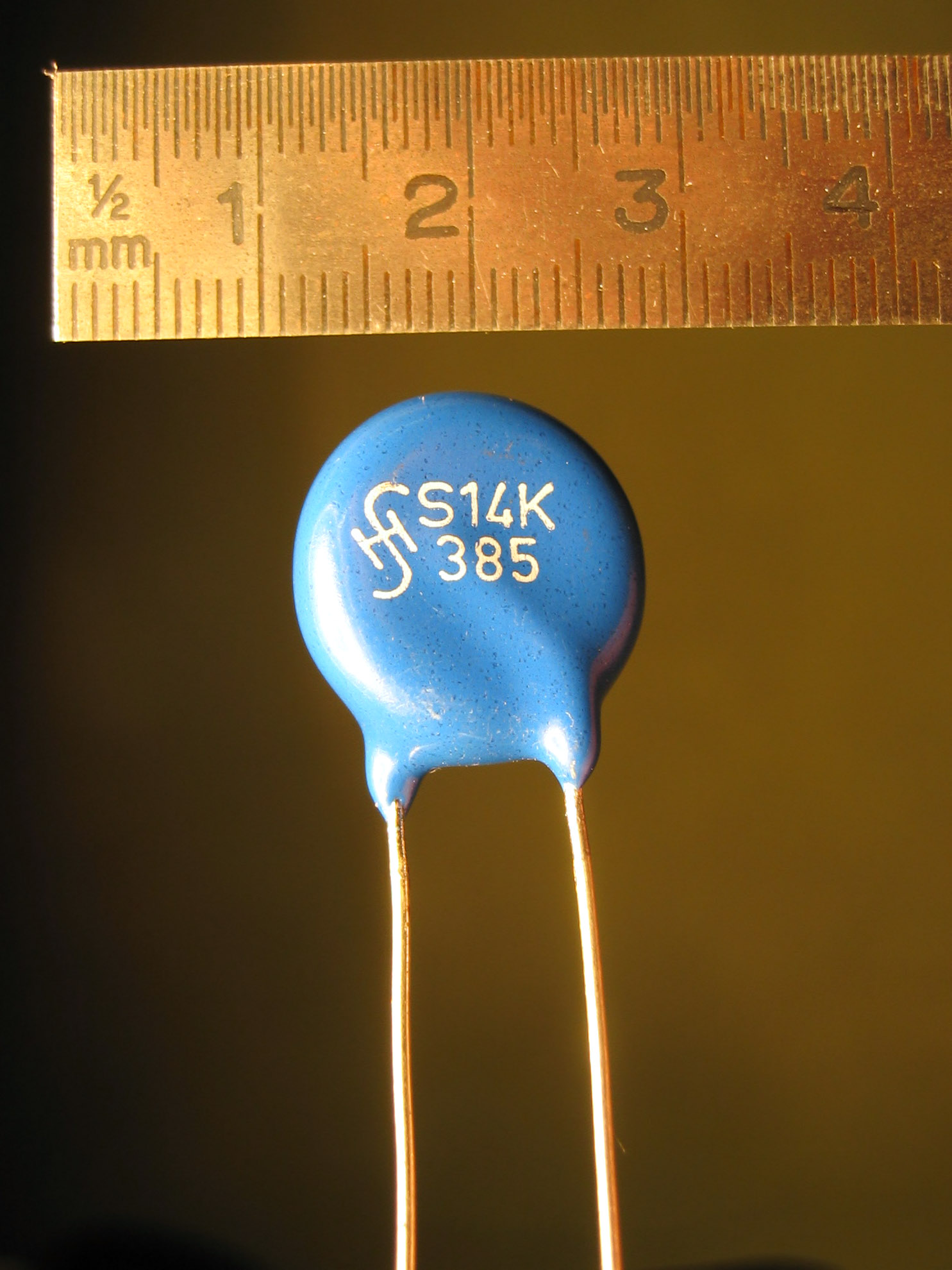 Figure 7 — A metal-oxide varistor (MOV): the classic blue radial disc, here a 14 mm "S14K385" rated for 385 V AC continuous. Beneath the epoxy is a sintered zinc-oxide ceramic; wired across the mai…